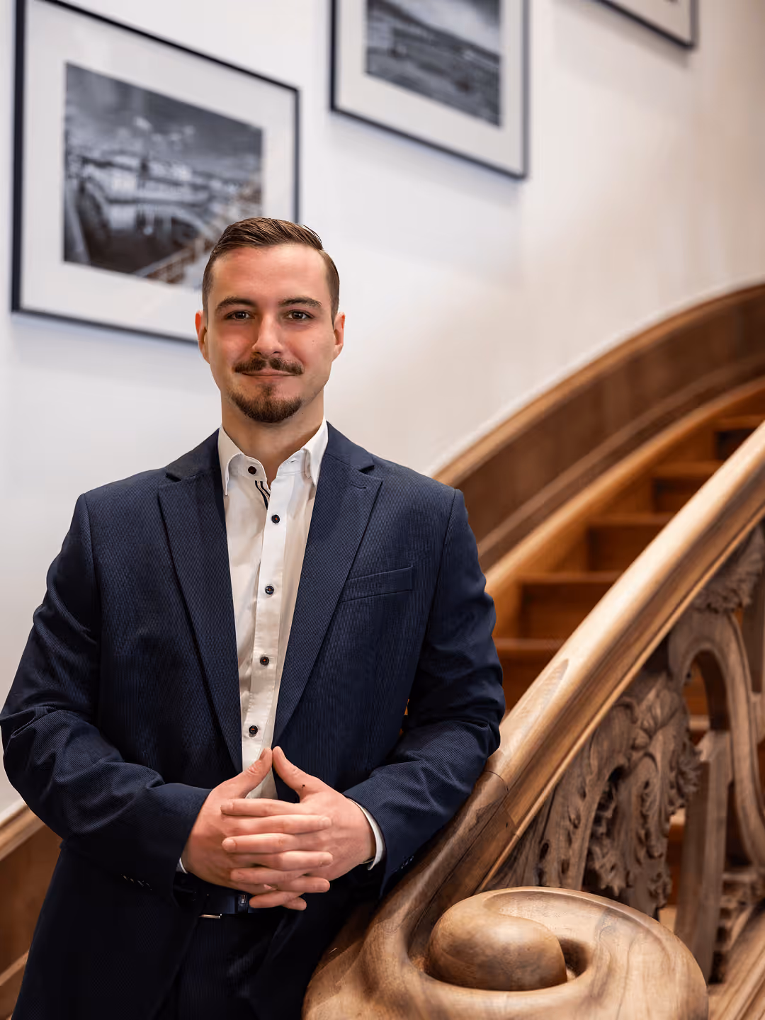 Mann im Anzug an geschnitztem Holzgeländer, lächelt leicht, Hintergrund mit gerahmten schwarz-weißen Fotos.