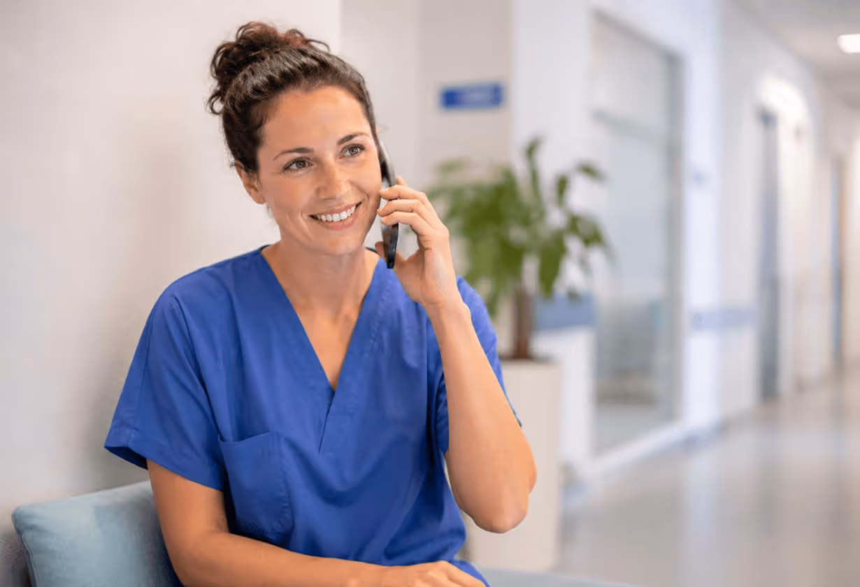 Krankenschwester in blauer Uniform telefoniert lächelnd im Krankenhausflur.