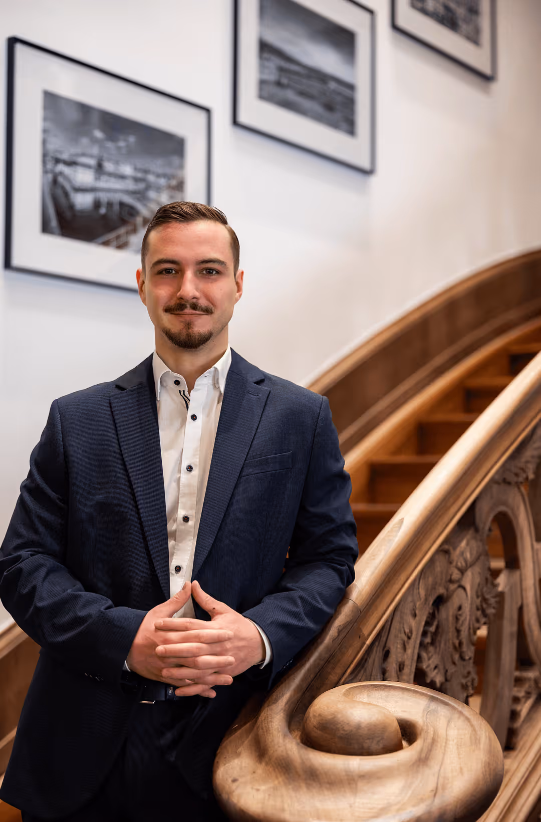 Mann im Anzug steht vor hölzerner Treppe, im Hintergrund gerahmte Schwarz-Weiß-Fotos an der Wand.